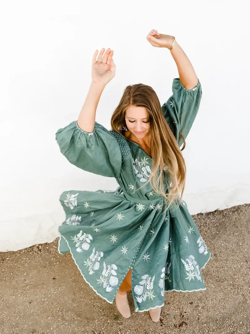 ElizaBeth Alexander, somatic coach, turning with both arms lifted in a green embroidered dress