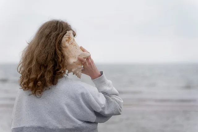 A person holding a large conch shell to their ear, facing a pale sea