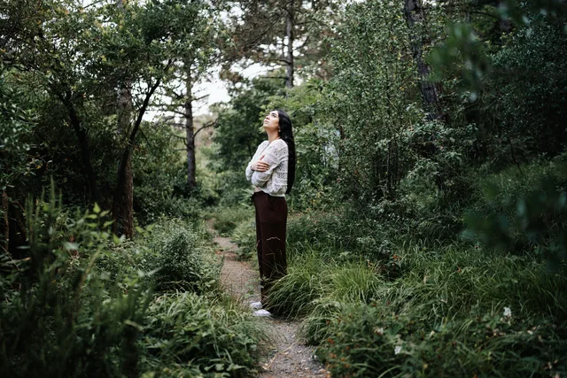 A woman standing alone on a woodland path, one hand resting on her chest, looking up through the trees